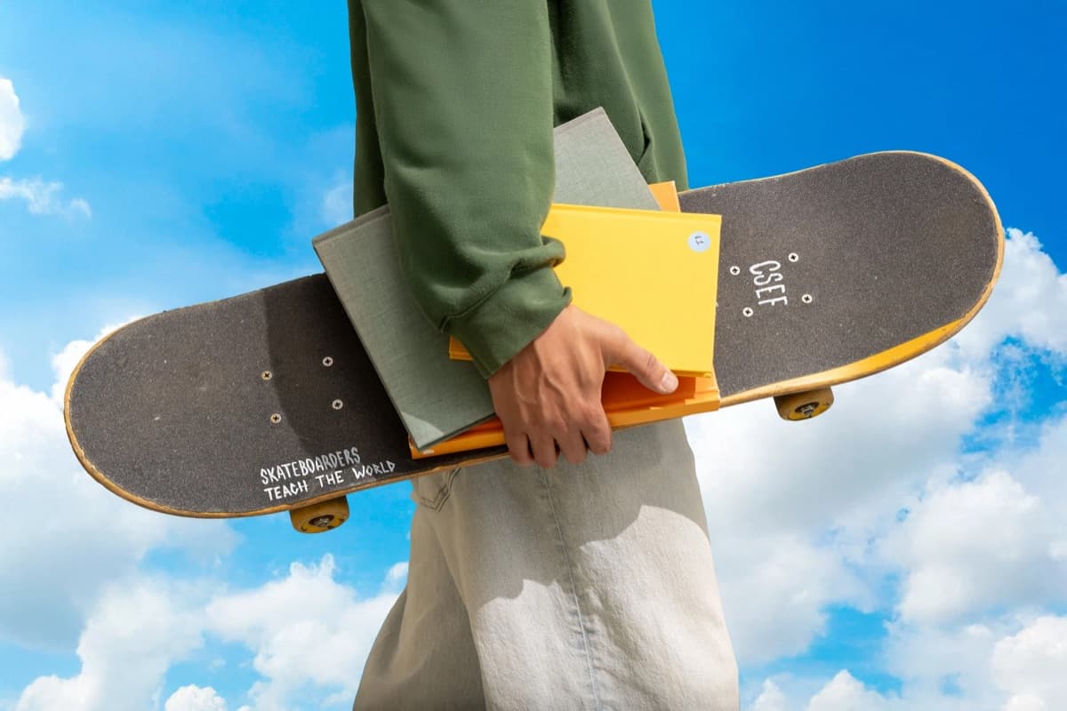 A person carrying a skateboard and a stack of notebooks under one arm, against a blue sky with clouds
