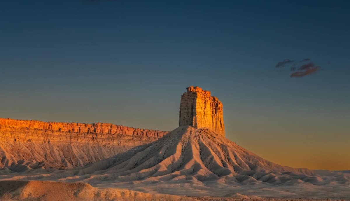 A lone sandstone butte rising above pale, eroded slopes at sunset, under a deep blue sky