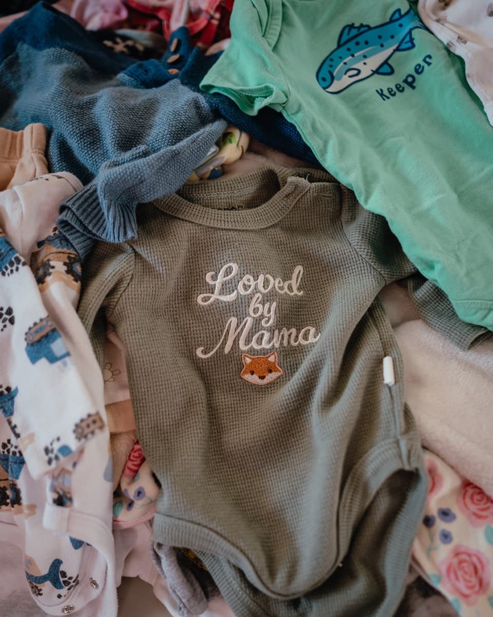 A pile of baby clothes, with a green onesie embroidered "Loved by Mama" on top